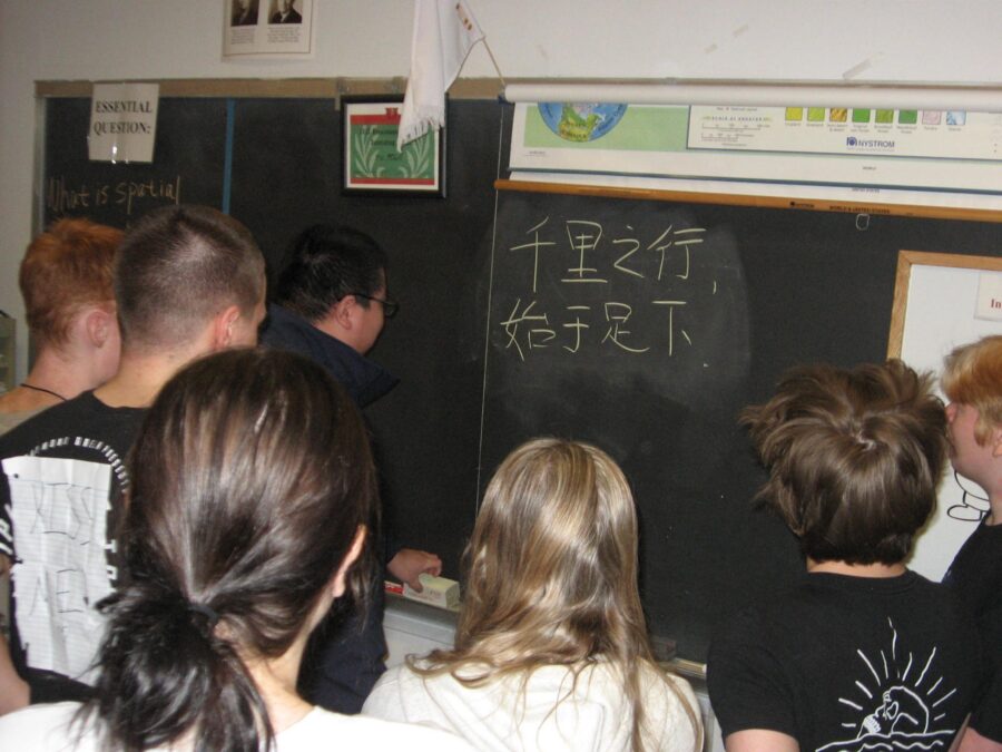 Mr. Fang teaching Chinese to students. The message reads, A thousand-mile trip starts with a single step.